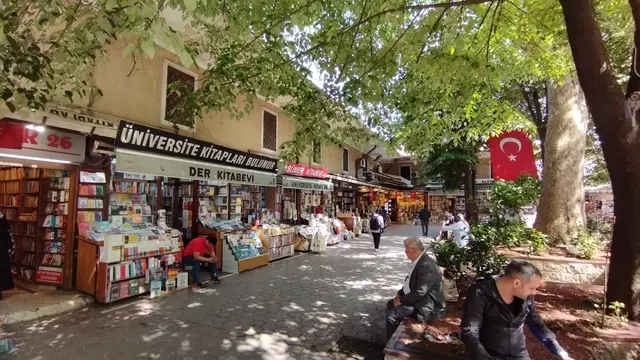 Fatih'te Sahaflar Çarşısı'nın avlusu; solda tabelalı kitapçı dükkânları ve döner sergi standları, sağda kalın gövdeli çınar ile daldan sarkan Türk bayrağı, zeminde altıgen parke.