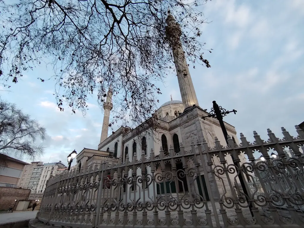 Pertevniyal Valide Sultan Camii'ne alttan bakış; demir parmaklıklı avlu duvarı, yapraksız ağaç dalları, minare ve kubbe.
