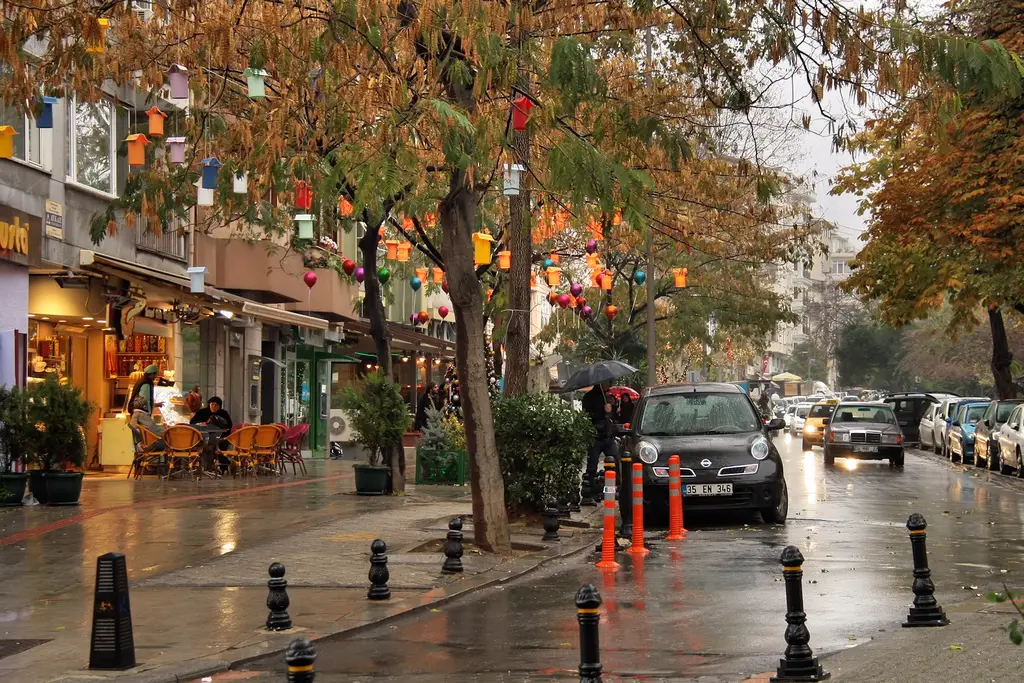 Moda'da yağmurlu bir sokak; sonbahar yapraklı ağaçlar, ıslak kaldırım, kafe masaları ve dizili babalar.