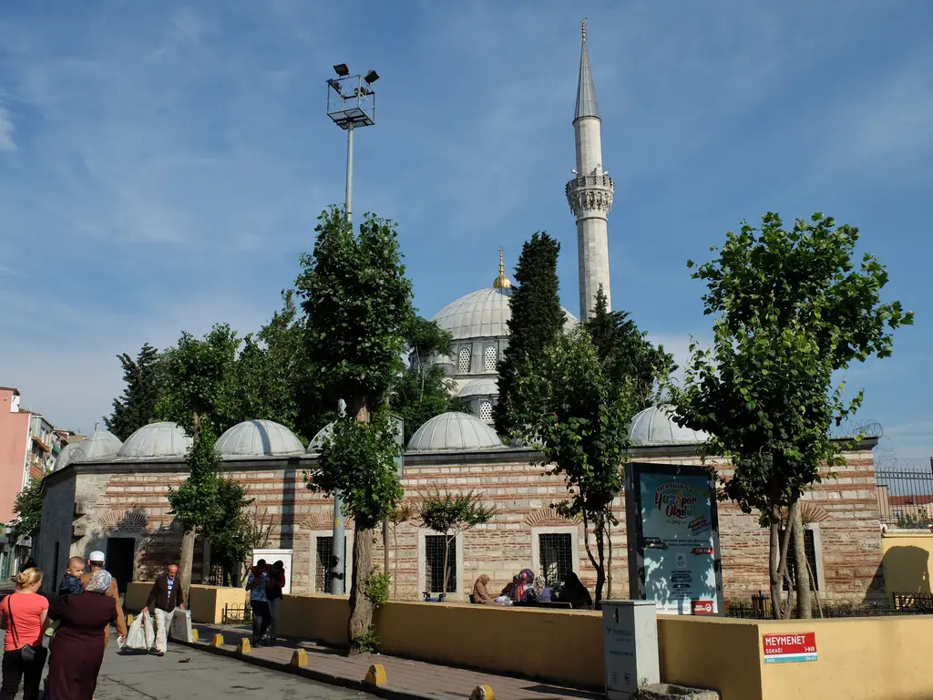 Caddeden Nişancı Mehmed Paşa Camii; taş ve tuğla avlu duvarı, üstünde kubbe dizisi, ağaçlar, minare ve kaldırımda yürüyen insanlar.
