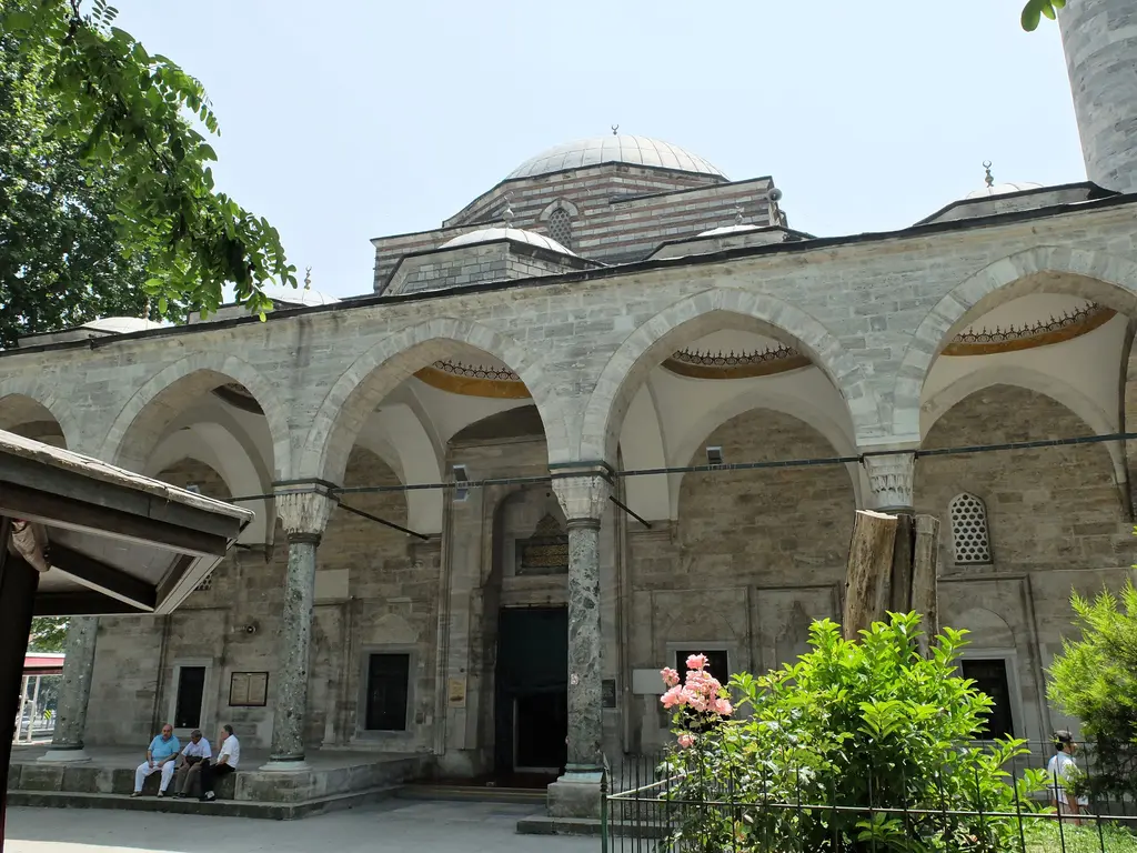 Murat Paşa Camii revakına yandan bakış; iki renkli taş kemerler, kubbeler, ağaç gölgesi ve önündeki bankta oturan insanlar.