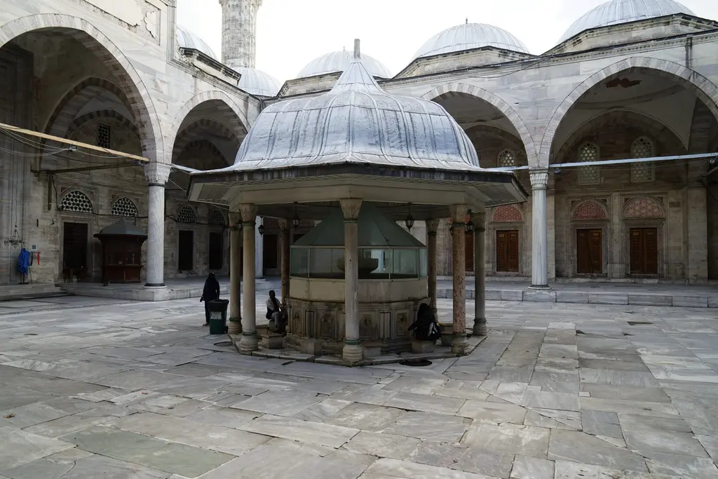 Şehzade Camii'nin şadırvanı zemin seviyesinden; yeşil şebekeli mermer gövde, kurşun kaplı külah, arkada revak kemerleri ile ahşap kapılar, ıslak mermer döşeme.