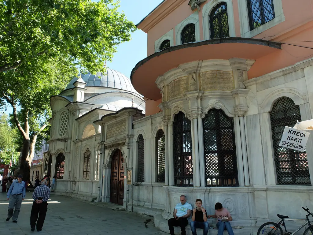 Eyüp'te Şah Sultan Külliyesi'nin cadde cephesi; şebekeli pencereli mermer türbe, çıkıntılı sebil ve önünde oturan insanlar.