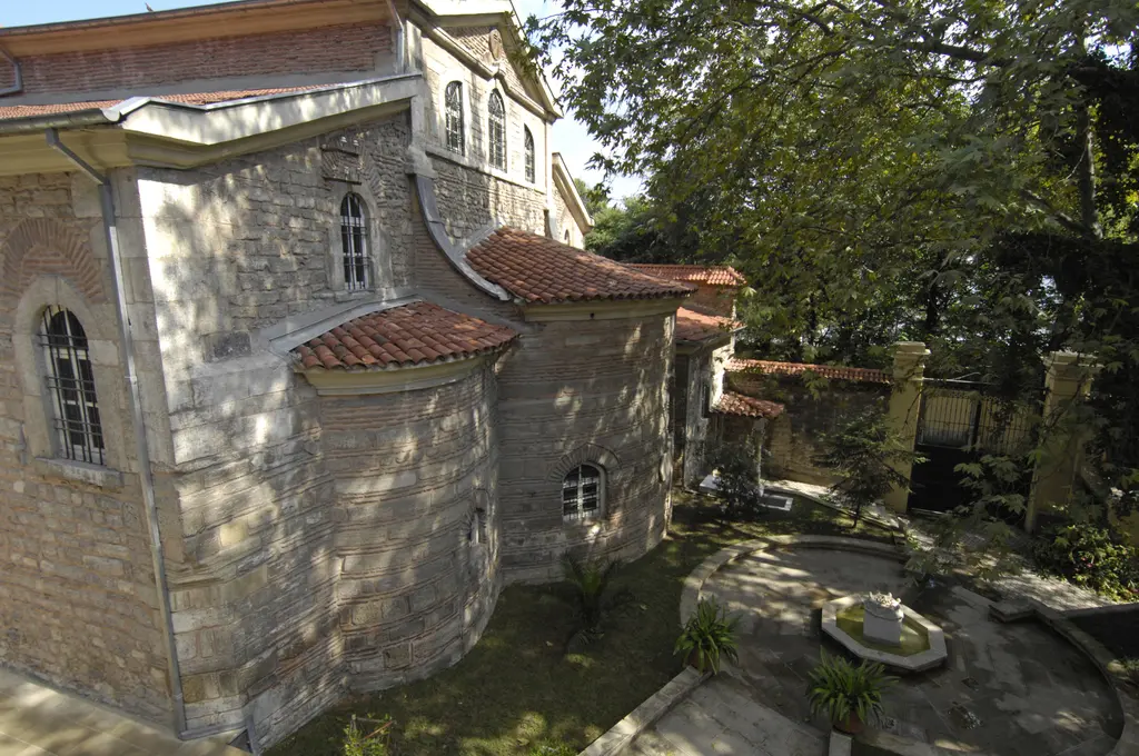 Aya Yorgi Kilisesi arkadan ve yandan; kiremit çatı katmanları, taş beden duvarları, bahçedeki mermer havuz ve gölge veren ağaçlar.