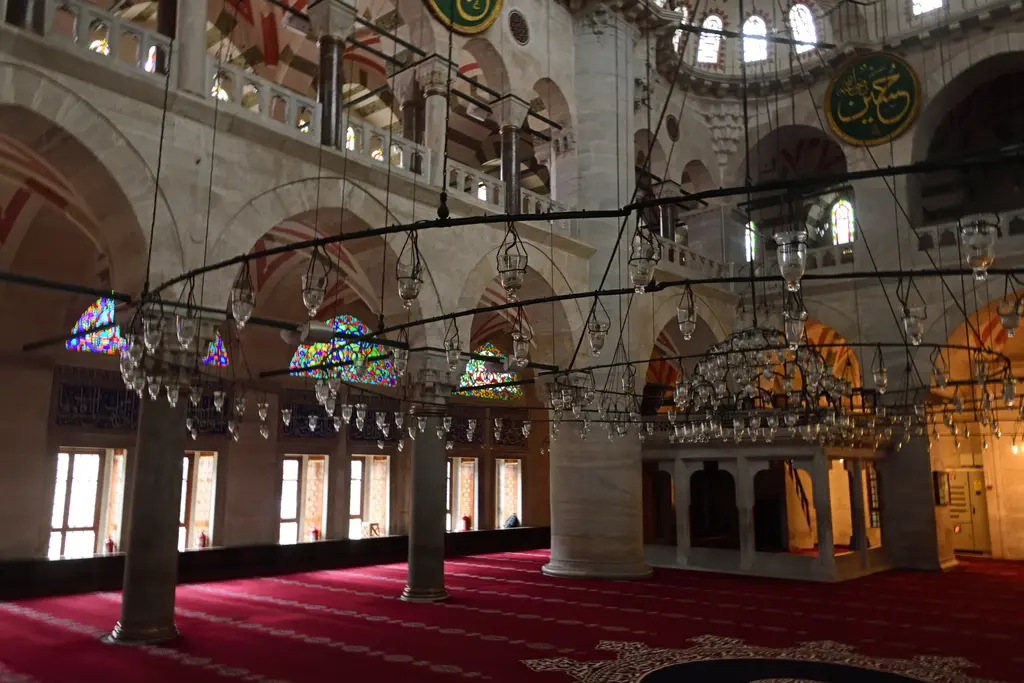 Kılıç Ali Paşa Camii'nin harimi; kırmızı halı, alçakta asılı avize çemberleri, ahşap mahfil ve kemerler arasında renkli camlı pencereler.