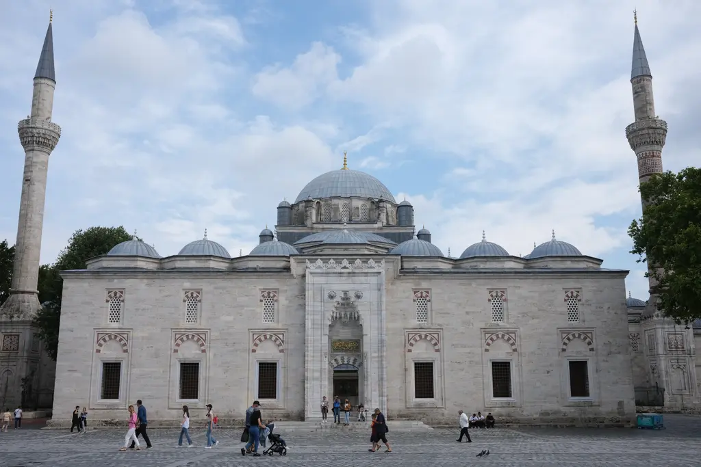 Beyazıt Camii önden; ortada kademelenen kubbeler, iki yanda birer ince minare, altta avlu duvarı ile mermer basamaklar ve meydanda yürüyen insanlar.
