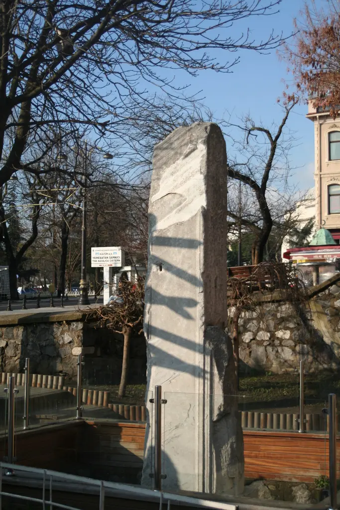 Kışın Milyon Taşı; cam korkuluklu çukurun içinde yükselen mermer ayak, arkada çıplak ağaç dalları, taş duvar ve mavi gök.