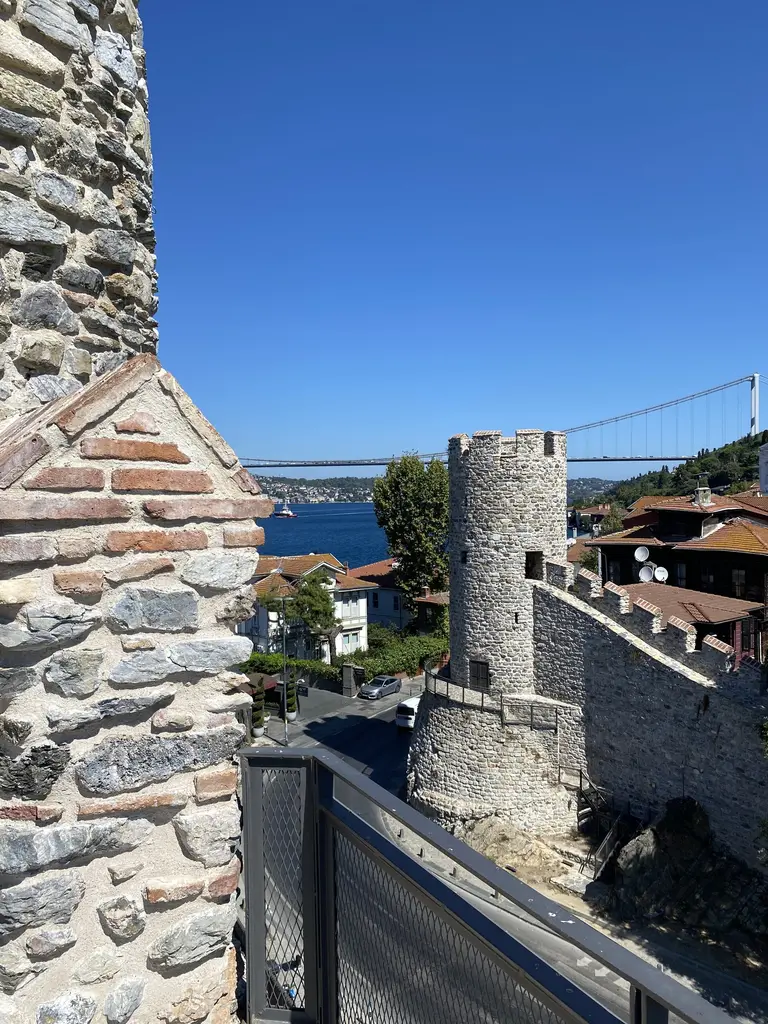 Anadolu Hisarı'nın içinden; taş duvar kenarından görünen yuvarlak kule, kırmızı çatılar ve uzakta köprü.