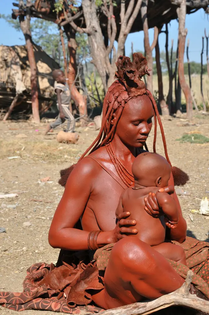 Namibya'da bir Himba kadını; kızıl aşı toprağıyla boyalı teni, örgülü saçları ve boncuk takılarıyla kucağında bebeğini tutuyor.