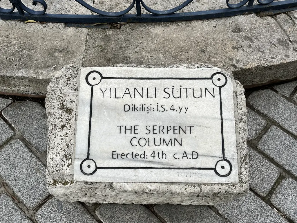 Yılanlı Sütun'un mermer tanıtım levhası; Türkçe ve İngilizce "Yılanlı Sütun, dikilişi İS 4. yüzyıl" yazısı, arkada parmaklık.