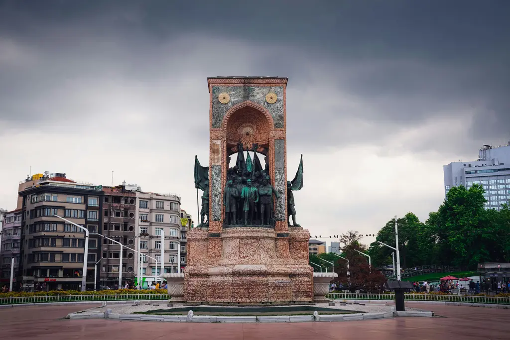 Kapalı bir günde Cumhuriyet Anıtı; tuğla renkli kemerli gövde, içindeki bronz figürler ve arkada koyu bulutlar.