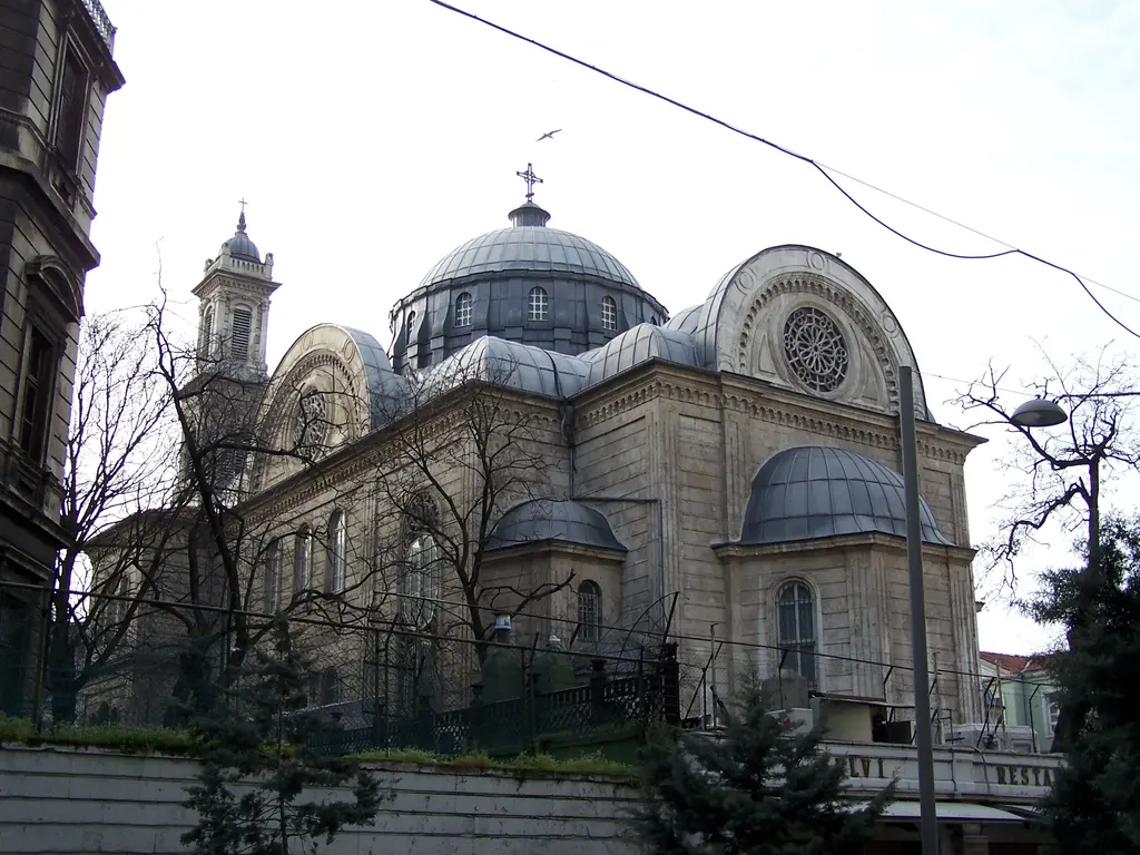 Aya Triada Kilisesi; bahçedeki ağaçların arasından görülen kubbe, çan kuleleri, yarım kubbeler ve önde demir parmaklık.