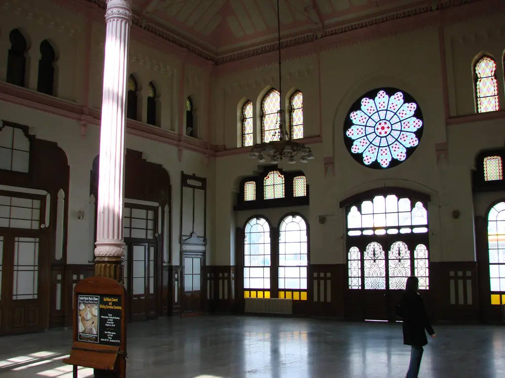 Sirkeci Garı'nın salonu diğer yönden; ortada yivli dökme demir sütun, sağda ışıyan gül pencere, gri zeminde yürüyen siyah paltolu bir kişi ve salonun ortasında danışma standı.