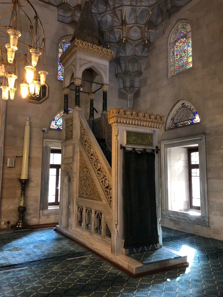 Şişli Camii'nin minberi; beyaz mermer korkuluk ve külah, yanında vitraylı pencereler ve önde mavi desenli halı.