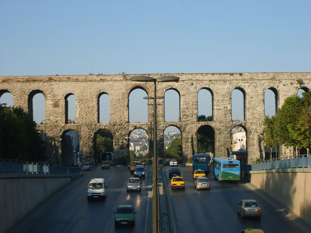 Bozdoğan Kemeri yakından, bulvarın üzerinde; kemer gözlerinden gökyüzü ile şehir silueti görünüyor, altta beyaz minibüsler, sarı taksiler ve sokak lambaları.