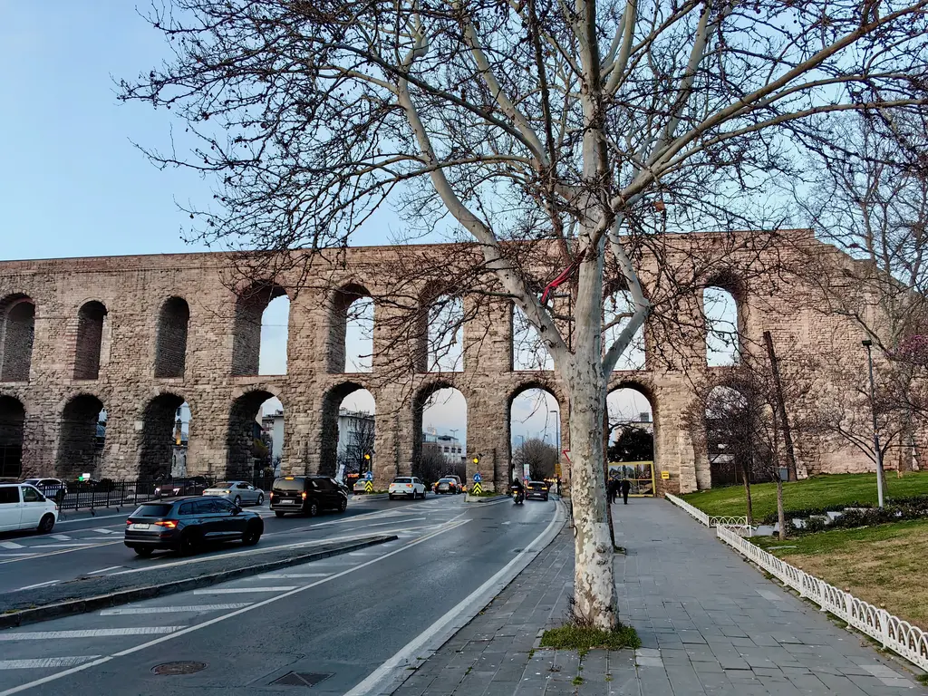 Bozdoğan Kemeri'nin önünde yapraksız kocaman bir çınar; dalları üst yarıyı ağ gibi kaplıyor, arkada iki katlı kemer dizisi, altta bulvar ve park etmiş koyu renk araçlar.