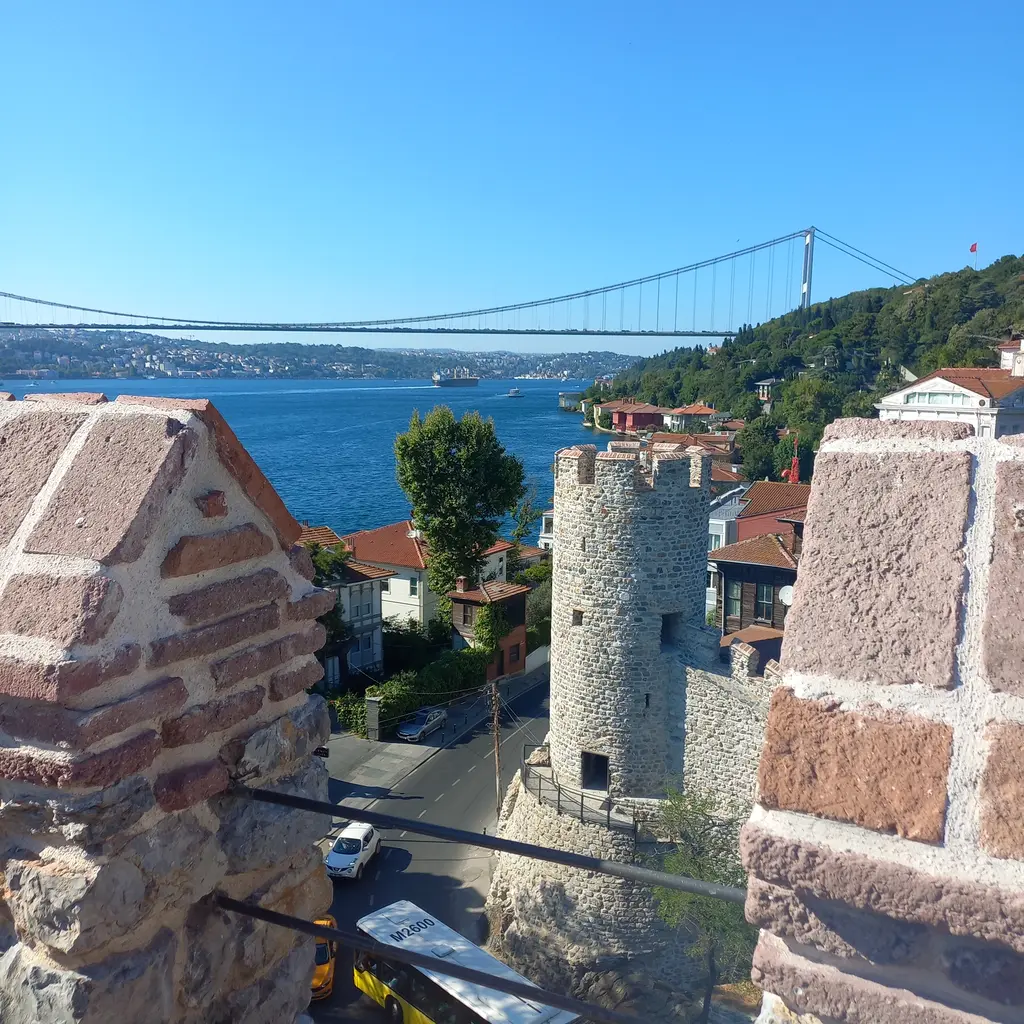 Anadolu Hisarı'nın iç kalesinden manzara; tuğla mazgalların arasından yuvarlak kule, yalılar ve uzakta asma köprü.