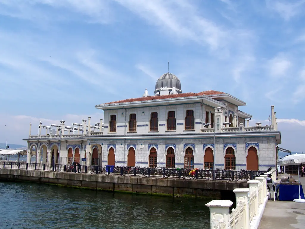 Adalar'da Büyükada iskelesi; kubbeli, sivri kemerli pencereli beyaz iskele binası, mavi deniz ve parçalı bulutlu gökyüzü.