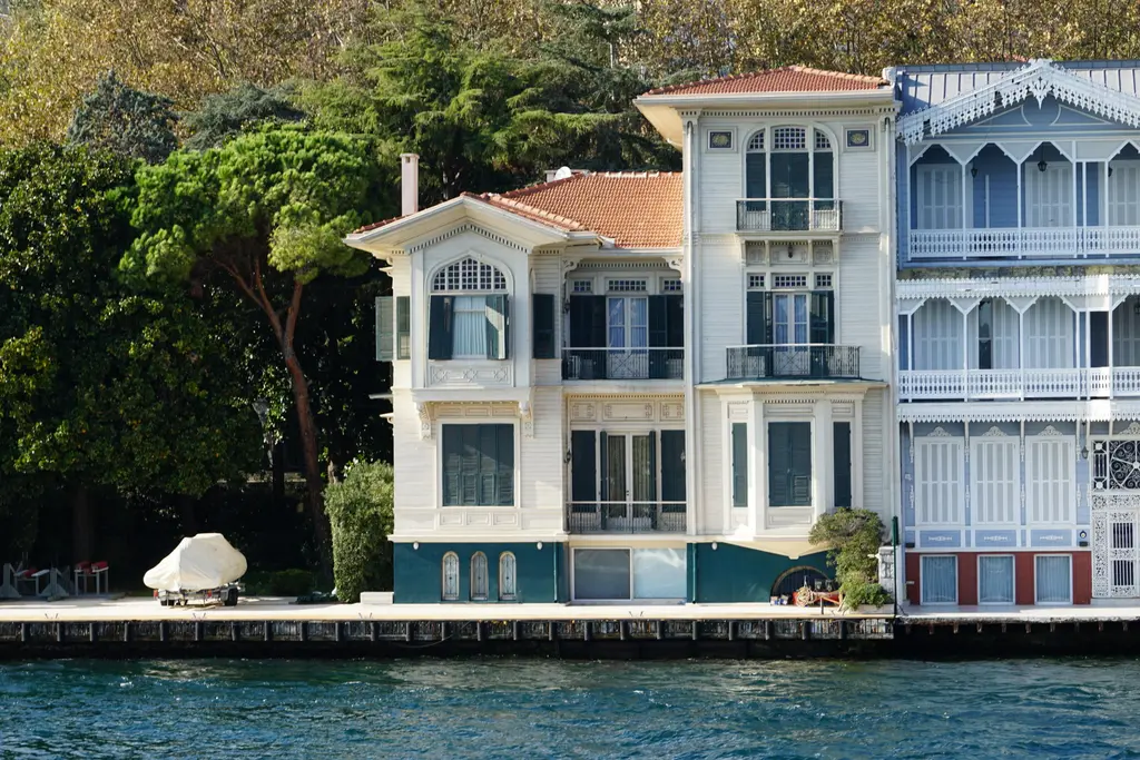 Gazioğlu Yalısı; beyaz ahşap cephe, kiremit çatı, mavi çerçeveli pencereler ve önde ağaçlarla rıhtım.