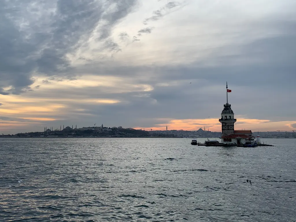 Salacak kıyısından Kız Kulesi; bulutlu gökyüzü, ufukta turuncu ışık, denizin ortasında kule ve kırmızı çatılar.