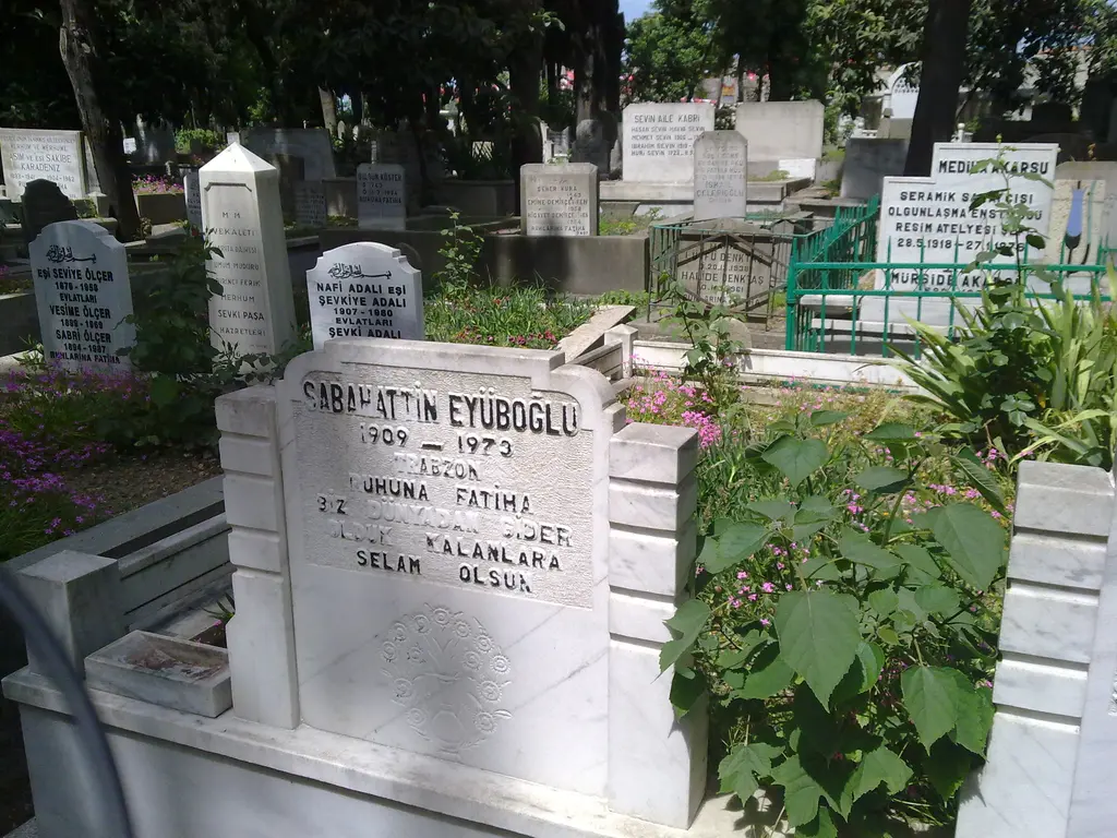 Beyaz mermer mezar taşı; üzerinde adların ve tarihlerin yazılı olduğu kitabe, çevresinde yeşillik ve başka mezar taşları.