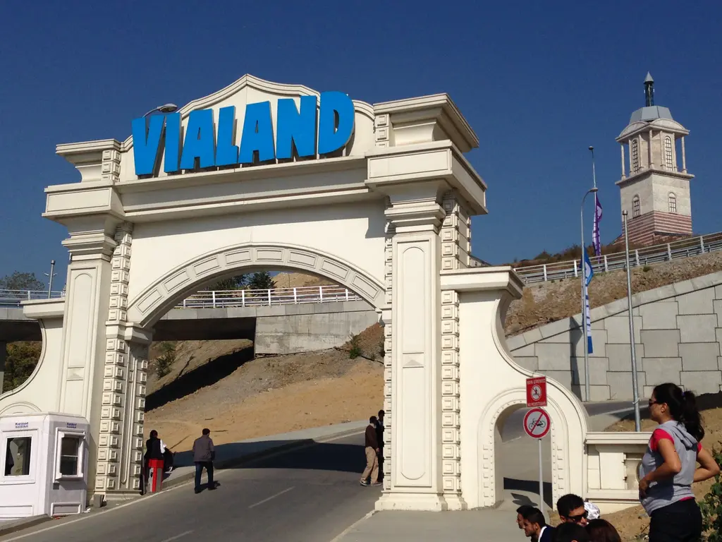 Vialand'in beyaz kemerli giriş kapısı; üstünde mavi harflerle "VIALAND" yazısı, arkada saat kuleli bir yapı ve kapıdan geçen ziyaretçiler.