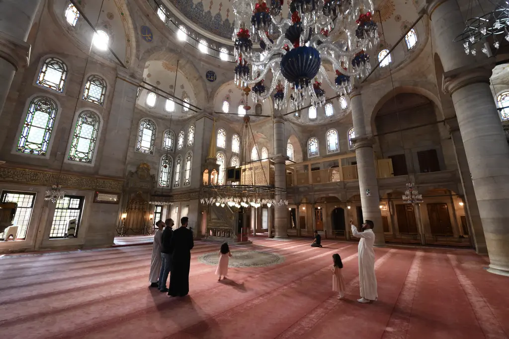 Eyüp Sultan Camii'nin harimi; kırmızı halı, kalın pâyeler, sarkan avize, iki kat pencere ve namaz kılanlar.