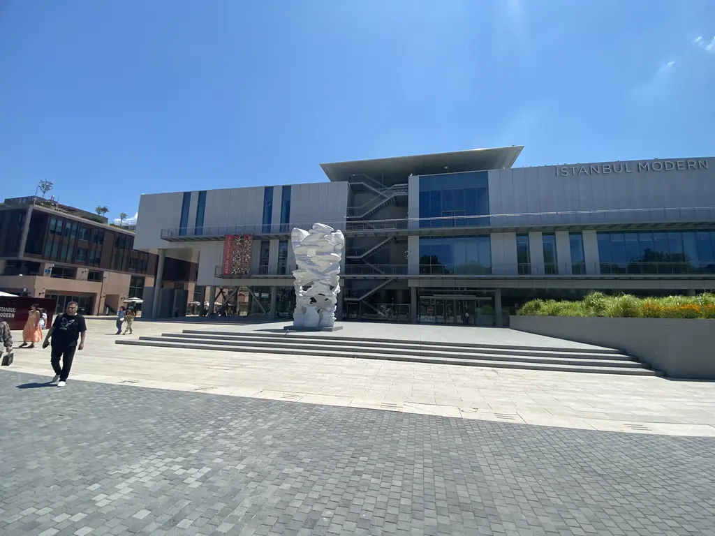Güneşli bir günde İstanbul Modern'in önü; meydanda beyaz bir heykel, basamaklar ve binanın gri cephesi.