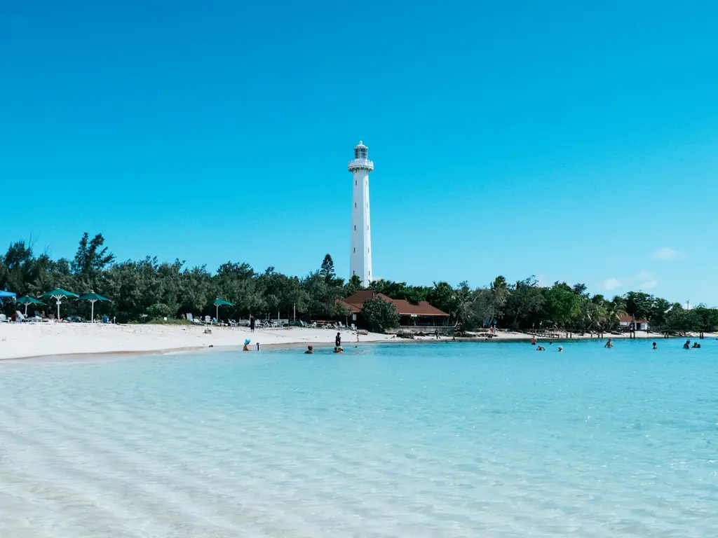 Yeni Kaledonya'da Nouméa açıklarındaki beyaz Amédée Deniz Feneri; önünde açık turkuaz suda yüzen insanlar ve ince kumlu plaj.