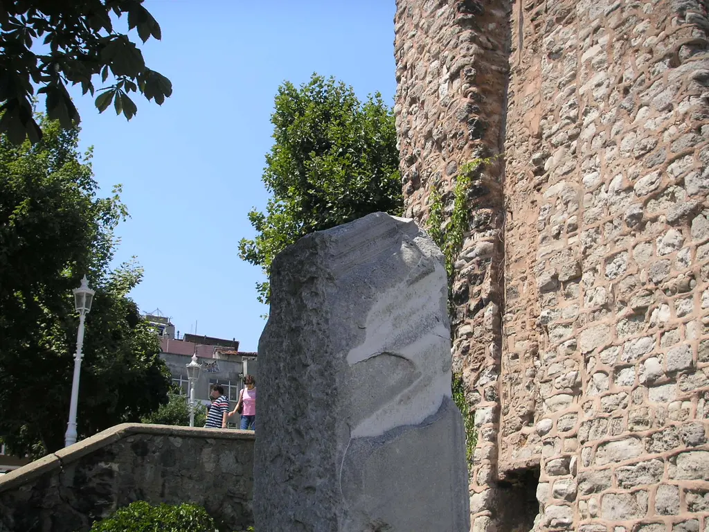 Milyon Taşı ve su terazisinin tuğla-taş karışık duvarı; önde mermer ayağın kırık üst ucu, arkada ağaçlar ve mavi gök.