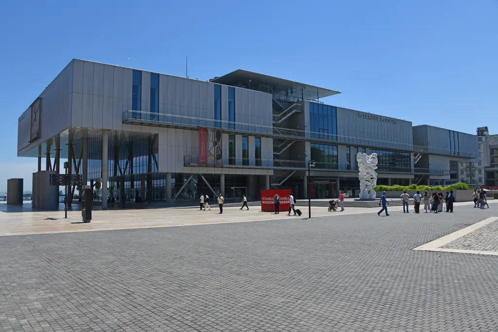 Açık bir günde İstanbul Modern ve önündeki geniş taş meydan; askıda duran kütleler, dış merdivenler ve yürüyen ziyaretçiler.