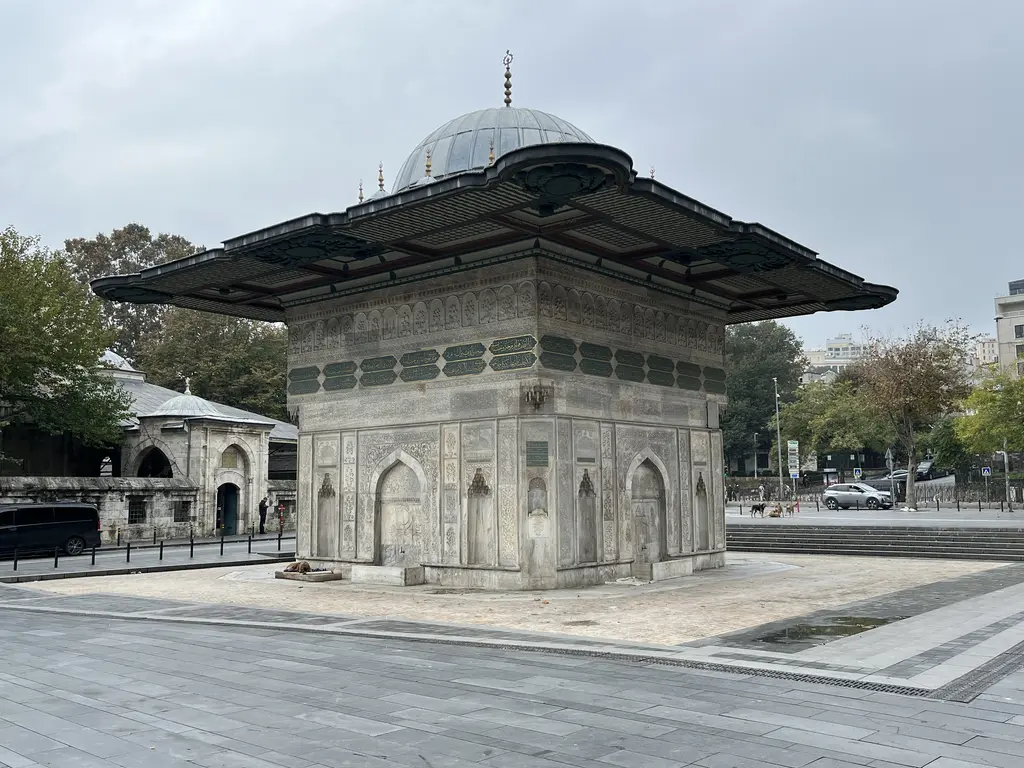 Tophane Çeşmesi; kare mermer gövde, yeşil zeminli yazı kuşağı, üstte geniş kurşun saçak ve tepede küçük kubbe, arkada ağaçlar.