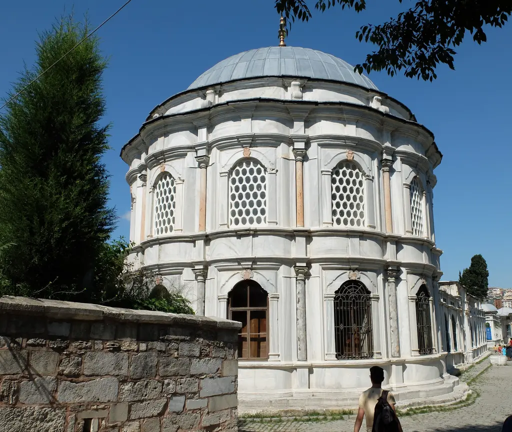 Mihrişah Valide Sultan Türbesi; beyaz mermer çok yüzlü gövde, iki sıra şebekeli pencere, kurşun kubbe, önünde servi ve taş duvar.
