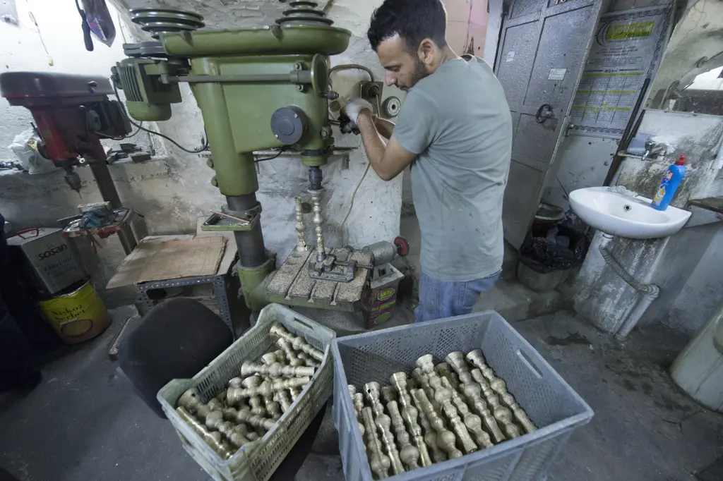 Büyük Valide Han'da bir atölye; yeşil gövdeli matkap tezgâhının başında çalışan genç bir adam, önde plastik kasalarda işlenmiş metal parçalar ve sağda lavabo.