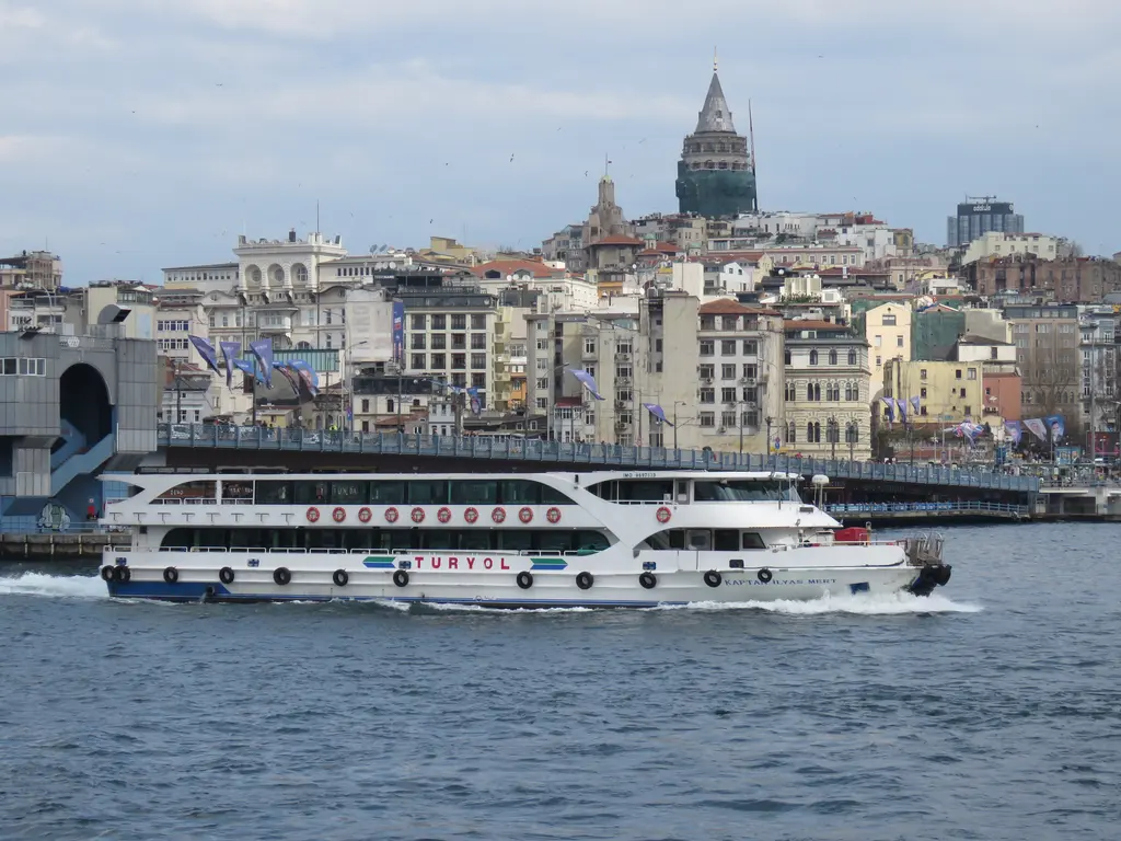 Köprünün önünden geçen beyaz şehir hatları vapuru; arkada köprü, Karaköy'ün yamaçları ve tepede Galata Kulesi.