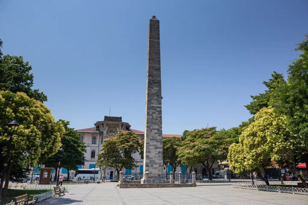 Sultanahmet Meydanı'nda Örme Sütun; kaba taş örgülü uzun gövde, çevresinde yeşil ağaçlar, arkada binalar ve açık mavi gökyüzü.