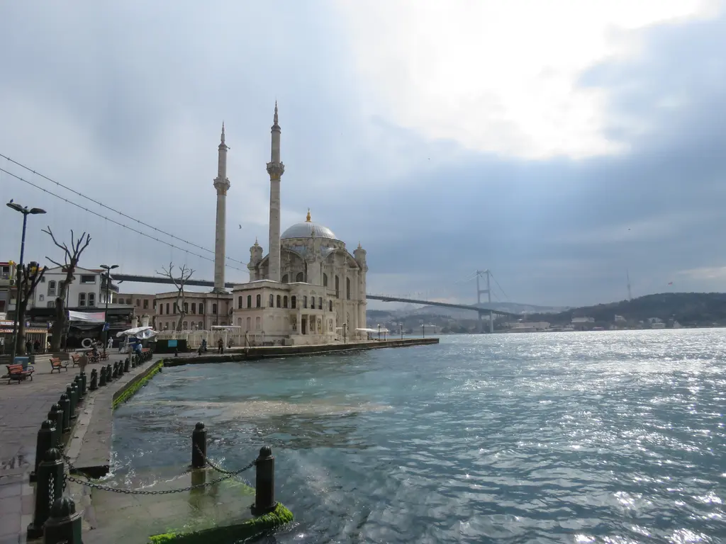 Kıyıdan Ortaköy Camii; önde parlayan Boğaz suyu ve rıhtım, ortada kubbeli cami ve minareleri, arkada köprü ve bulutlar.