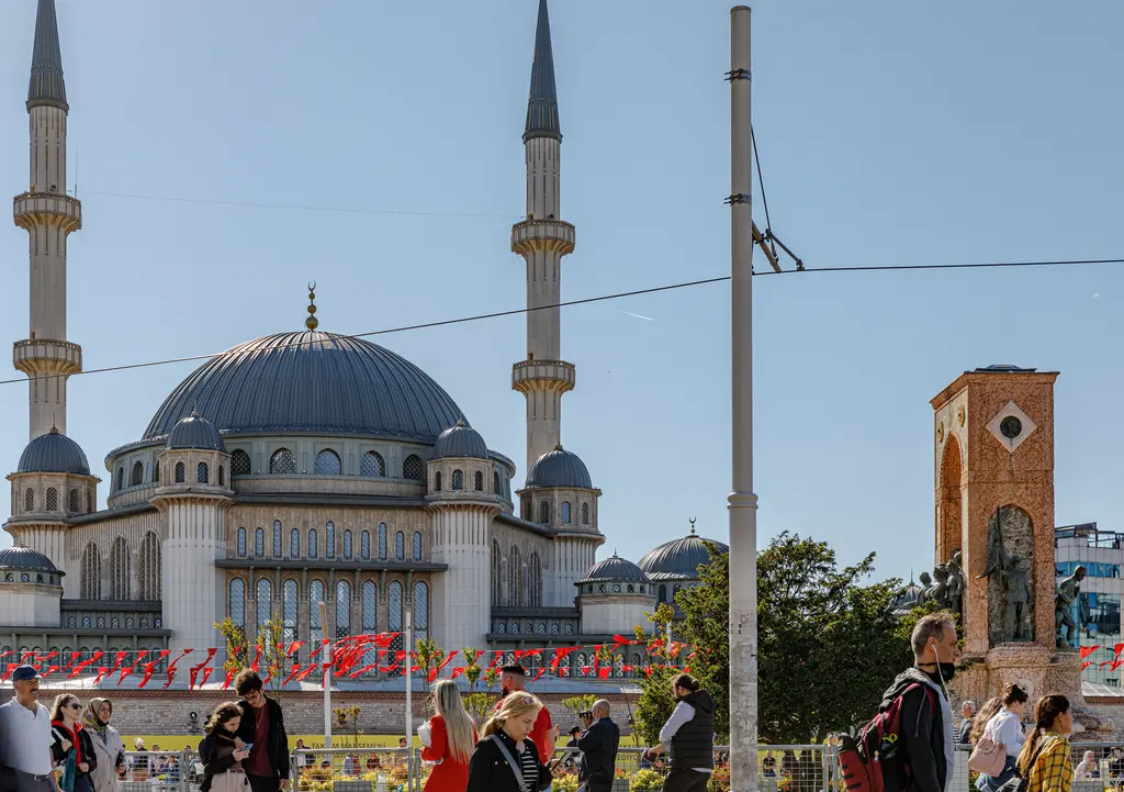 Taksim Camii'nin kubbesi ve iki minaresi yakından; sağda Cumhuriyet Anıtı'nın tuğla renkli gövdesi, önde bayraklar ve kalabalık.