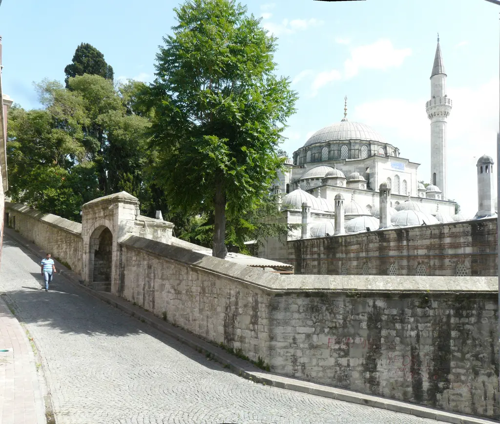 Kadırga'da yokuştan Sokullu Mehmed Paşa Camii; yol boyunca uzanan yüksek taş duvar, ağaçların arkasında kubbe ile minare ve eğimli kaldırım.