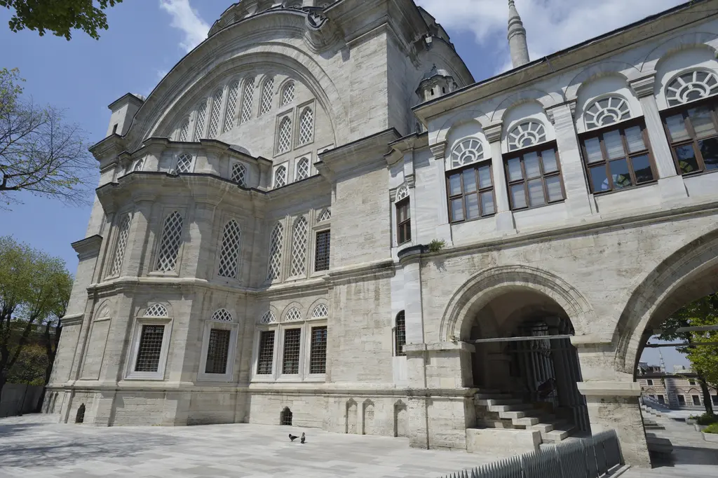 Nuruosmaniye Camii'nin doğu cephesi çapraz açıdan; büyük kemer, üst üste dizilen kemerli pencere sıraları, sağda revaklı kanat ve önde geniş mermer zemin.