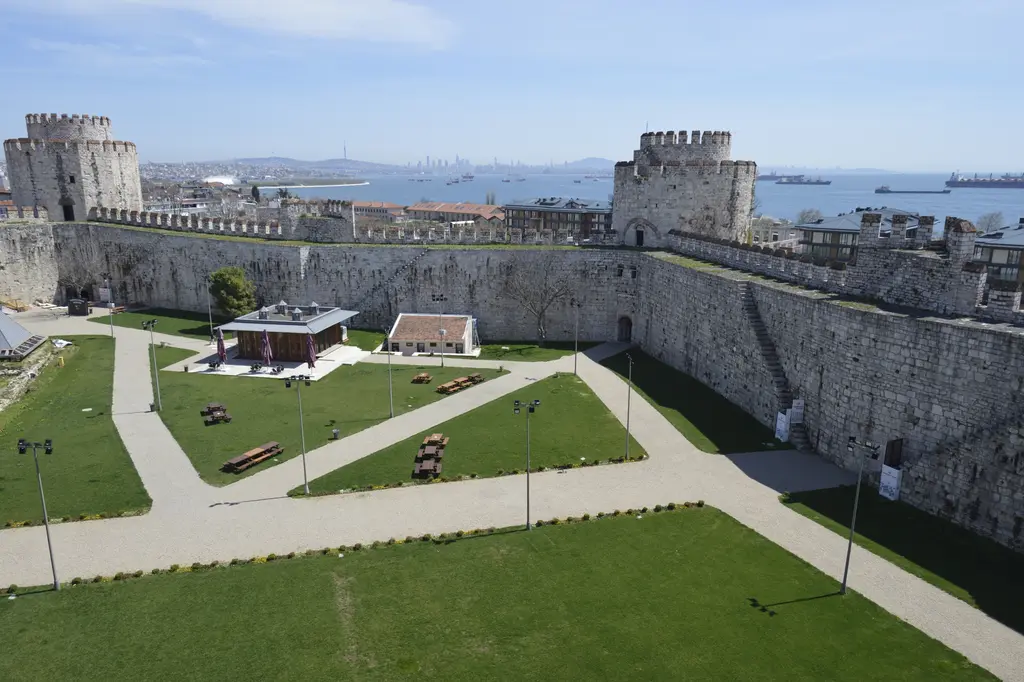 Yedikule Hisarı'nın avlusu duvarın üstünden; çimenlik alanda çapraz patikalar, piknik masaları, iki küçük yapı, sağda içeri inen taş merdiven ve uzakta Marmara Denizi.