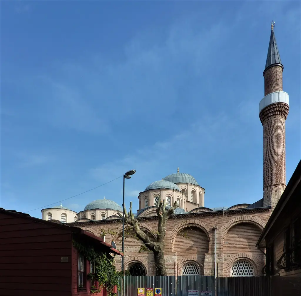 Zeyrek Camii'nin tek şerefeli ince minaresi; arkasında kurşun kaplı kubbeler ile tuğla gövde, önde eski ahşap evlerin çatıları, üstte bulutsuz mavi gökyüzü.