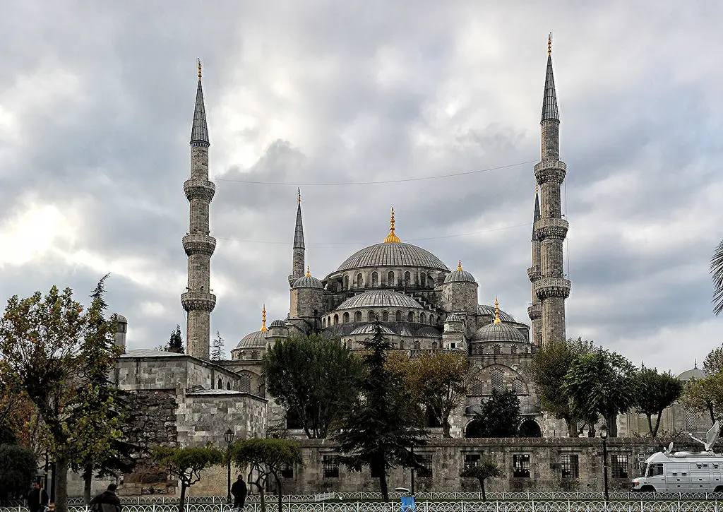 Sultanahmet Camii; altı minare, kademeli yükselen kubbeler, önde ağaçlar ve bahçe duvarı, üstte bulutlu gökyüzü.