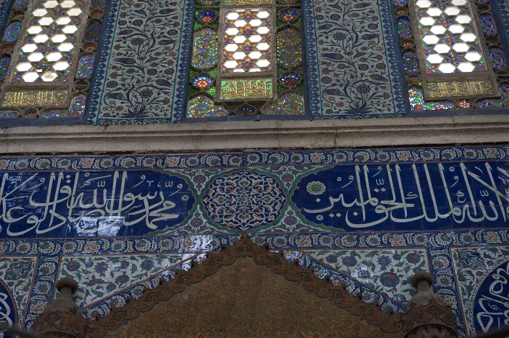 Sokullu Mehmed Paşa Camii'nde mihrap duvarı; renkli camlı pencereler arasında çiçek desenli çini panolar, altta lacivert zeminli beyaz hat kuşağı.