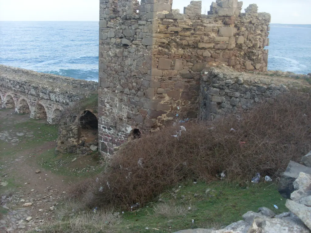 Rumeli Feneri Kalesi'nde burç; mazgallı taş kule, yanında kemerli sur duvarı, otlarla kaplı avlu ve arkada deniz.