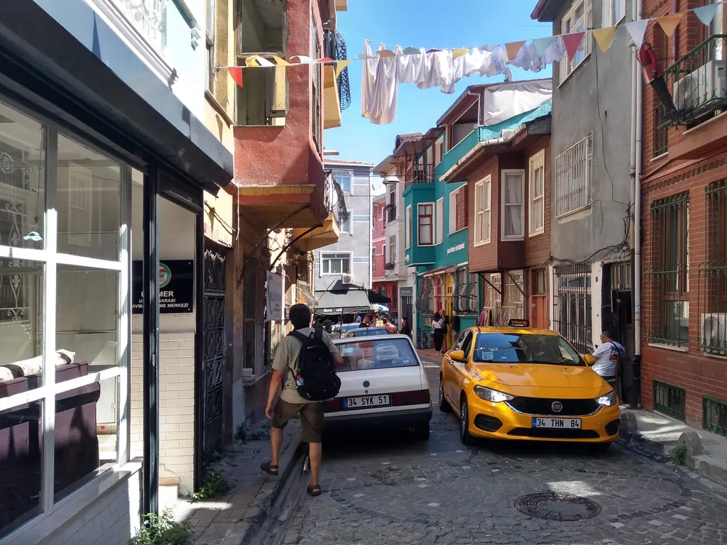 Balat'ta flamalarla süslü dar sokak; renkli cumbalı evler, ipte çamaşırlar, sarı taksi ve yürüyen insanlar.