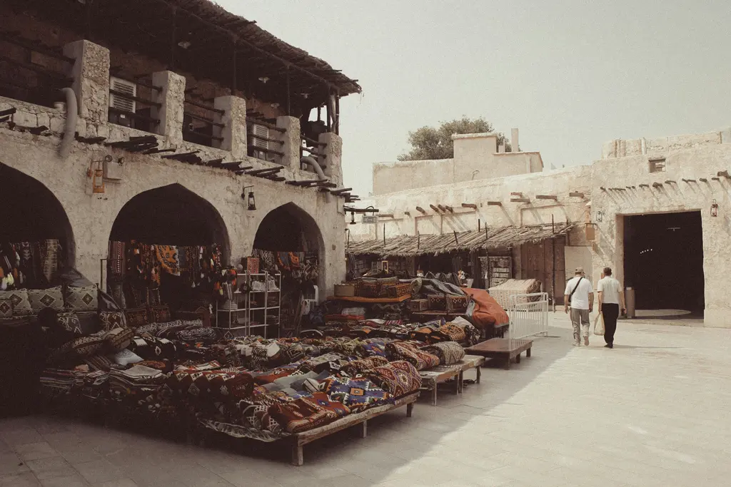 Souq Waqif'te üstü açık bir çarşı sokağı; kemerli arkadın altında sergilenen sepetler ve dokumalar, taş kaplı zemin ve gölgede yürüyen iki kişi.