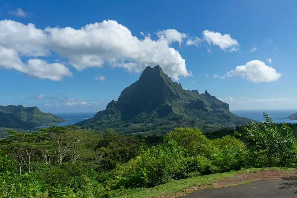 Moorea'nın iç kesiminde yükselen sivri volkanik zirve; zirveye tutunan beyaz bulutlar, etekleri saran tropik orman ve uzakta lagünün mavi suları.