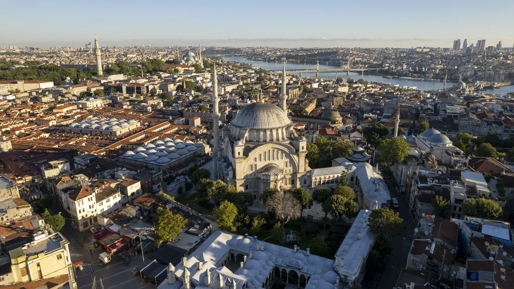 Nuruosmaniye Camii havadan; ortada caminin kubbesi ile iki minaresi, solda Kapalıçarşı'nın kurşun kaplı çatı denizi, arkada Haliç ve köprüler, sabah ışığı.