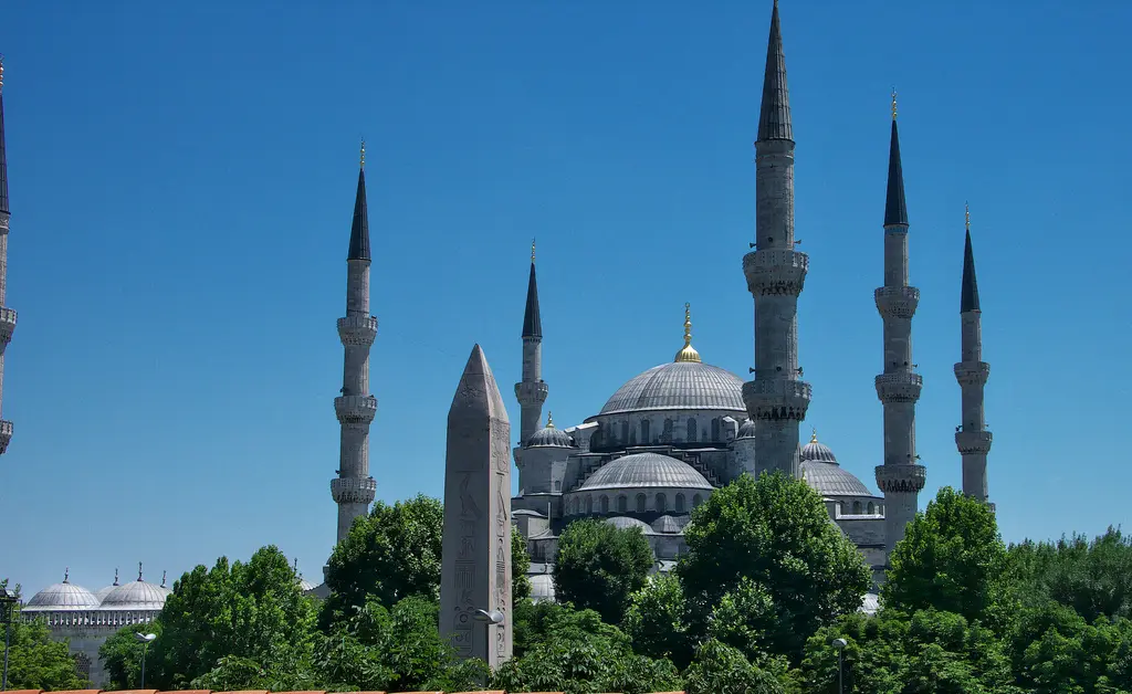 Dikilitaş ile Sultanahmet Camii; önde yeşil ağaçlar, ortada granit obelisk, arkada caminin kubbeleri ve altı minaresi, açık mavi gök.